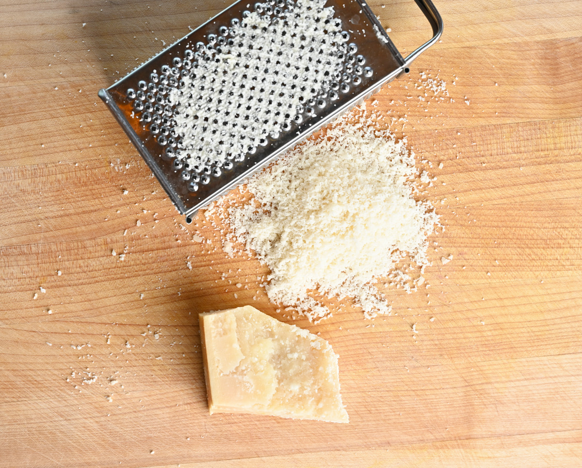 a pile of grated Parmigiano Reggiano sitting beside a metal grater and a block of cheese on a wooden cutting board