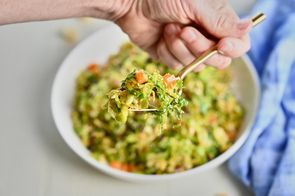 close up of Shredded Brussel Sprout Salad with miso on a gold fork over a white bowl with a blue cloth napkin beside it