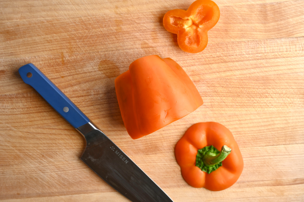 Pepper with the top cut off it laying on its side next to a chef's knife on a wooden cutting board