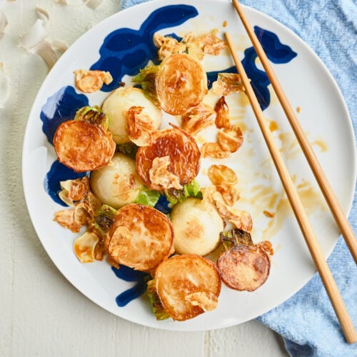 Half-eaten plate of turnips with chopsticks on a white plate with a blue accent, styled with a gold spoon, blue linen, and garlic peels.