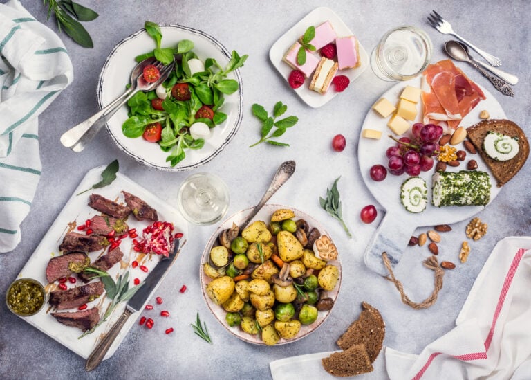 Dinner plated buffet style featuring a meat and cheese board, steak with chimichurri and pomegranate, roasted potatoes, and dessert, complemented by scattered herbs.
