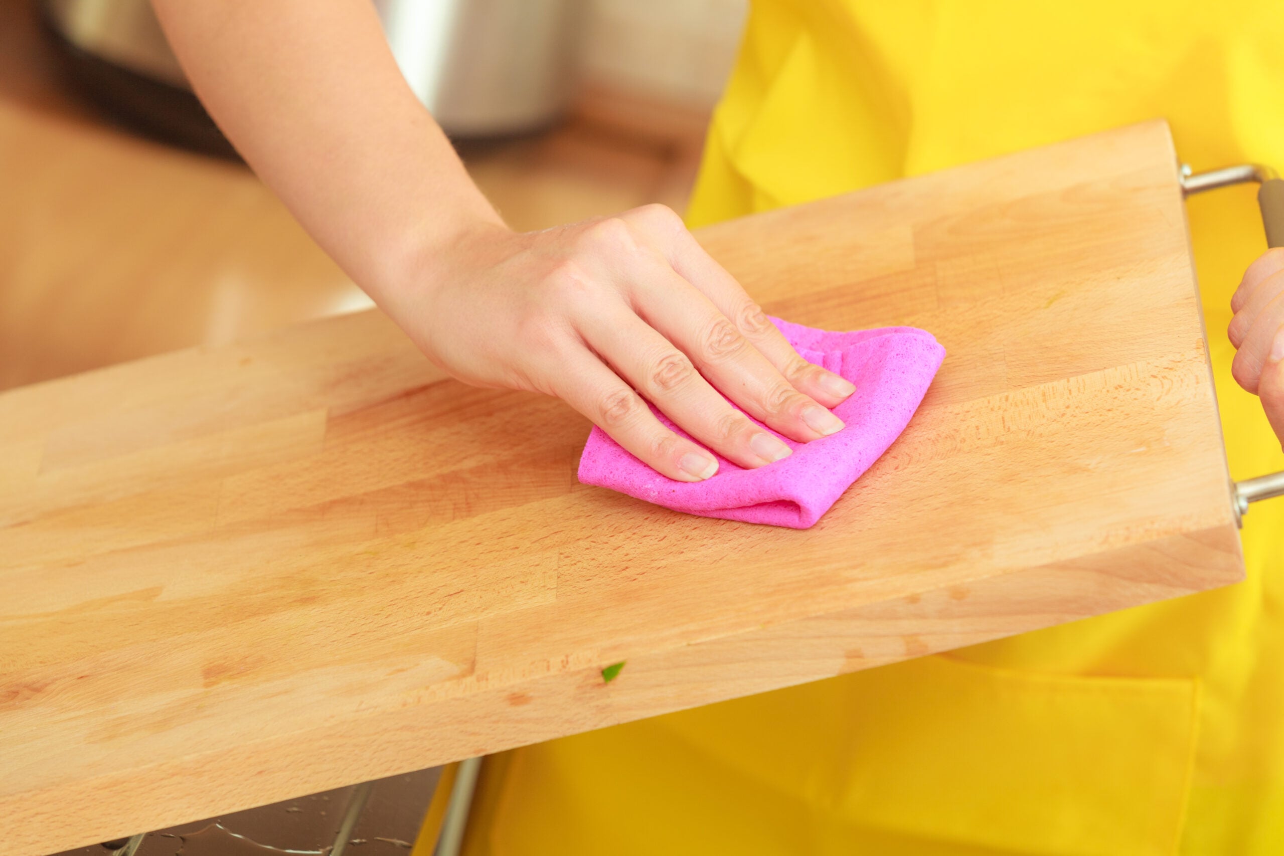 Woman in a yellow apron cleaning a cutting board with a pink sponge.