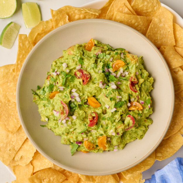 Overhead shot of finished guacamole in a bowl on a platter with tortilla chips, blue linen, limes, and cilantro.
