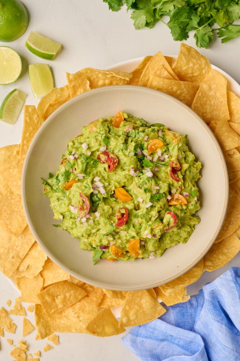 Overhead shot of finished guacamole in a bowl on a platter with tortilla chips, blue linen, limes, and cilantro.