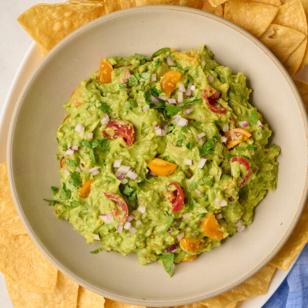 Close-up shot of finished guacamole in a bowl served with tortilla chips, styled with blue linen and limes.
