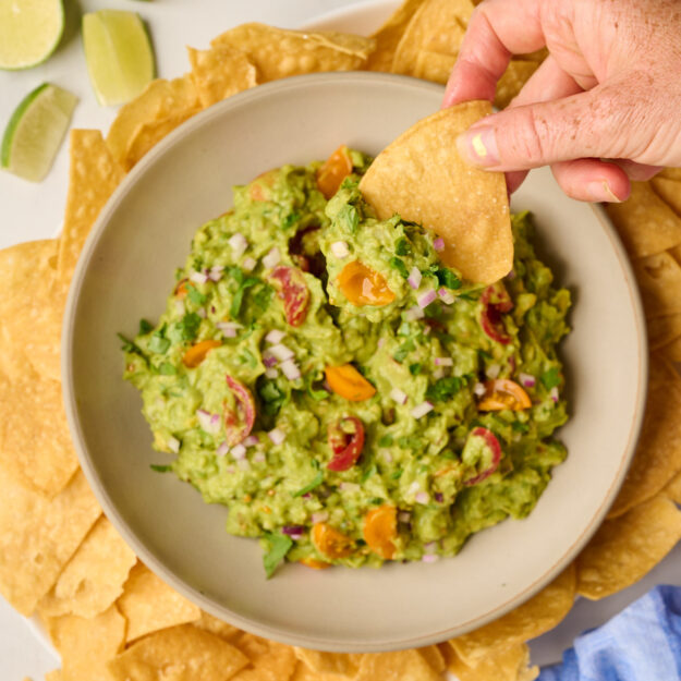 Hand scooping guacamole with a tortilla chip, with tortilla chips, limes, cilantro, and blue linen in the background.