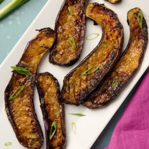 Plated miso eggplant garnished with thinly sliced green onions on a blue surface with pink linen and fresh green onions in the background.