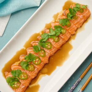 Fully plated salmon sashimi dressed with yuzu soy sauce, garnished with thinly sliced jalapeño and cilantro, styled with blue linen and chopsticks.