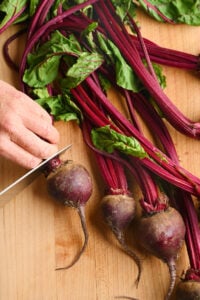 Hand slicing beet bulbs from greens on a cutting board.