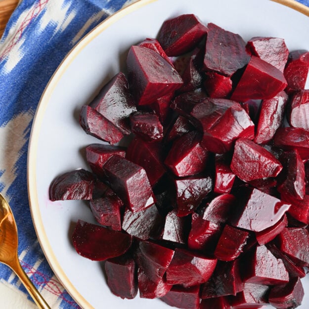 Close-up shot of roasted beets in a bowl with a gold spoon, blue ikat napkin, and wooden serving board.