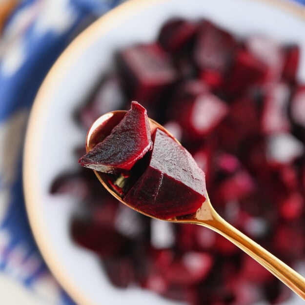 Gold spoon holding roasted beets over a finished plate of beets, ikat blue napkin, and wooden serving board.