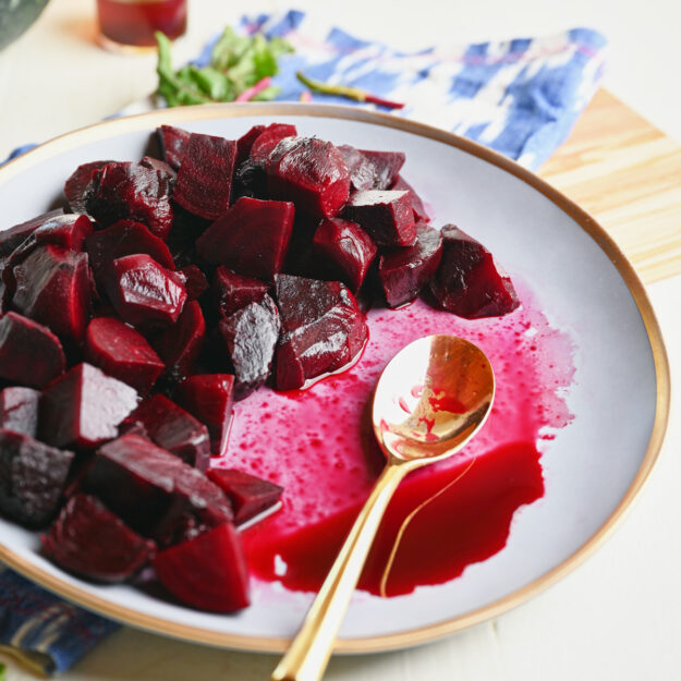 Side shot of a half-eaten plate of roasted beets with a gold spoon on a blue ikat napkin and wooden serving board.
