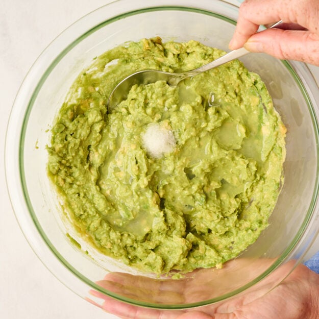 Hand holding a spoon in mashed avocado with salt and lime juice added, with a blue cutting board featuring diced tomatoes, jalapeño, red onion, and limes in the background.