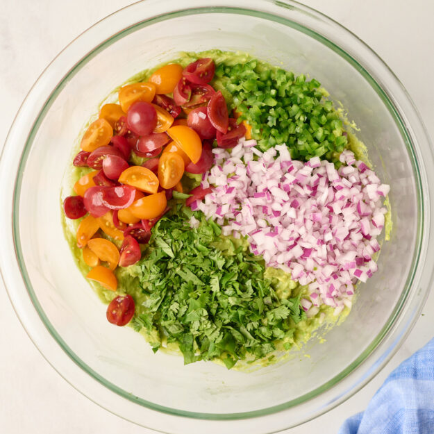 Glass bowl of mashed avocado topped with diced tomatoes, jalapenos, red onion, and cilantro, styled with blue linen, limes, and fresh cilantro in the background.