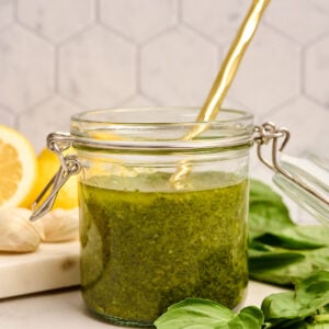 Finished jar of basil vinaigrette with a gold spoon inside, surrounded by fresh basil leaves, garlic, and lemon on a marble cutting board.