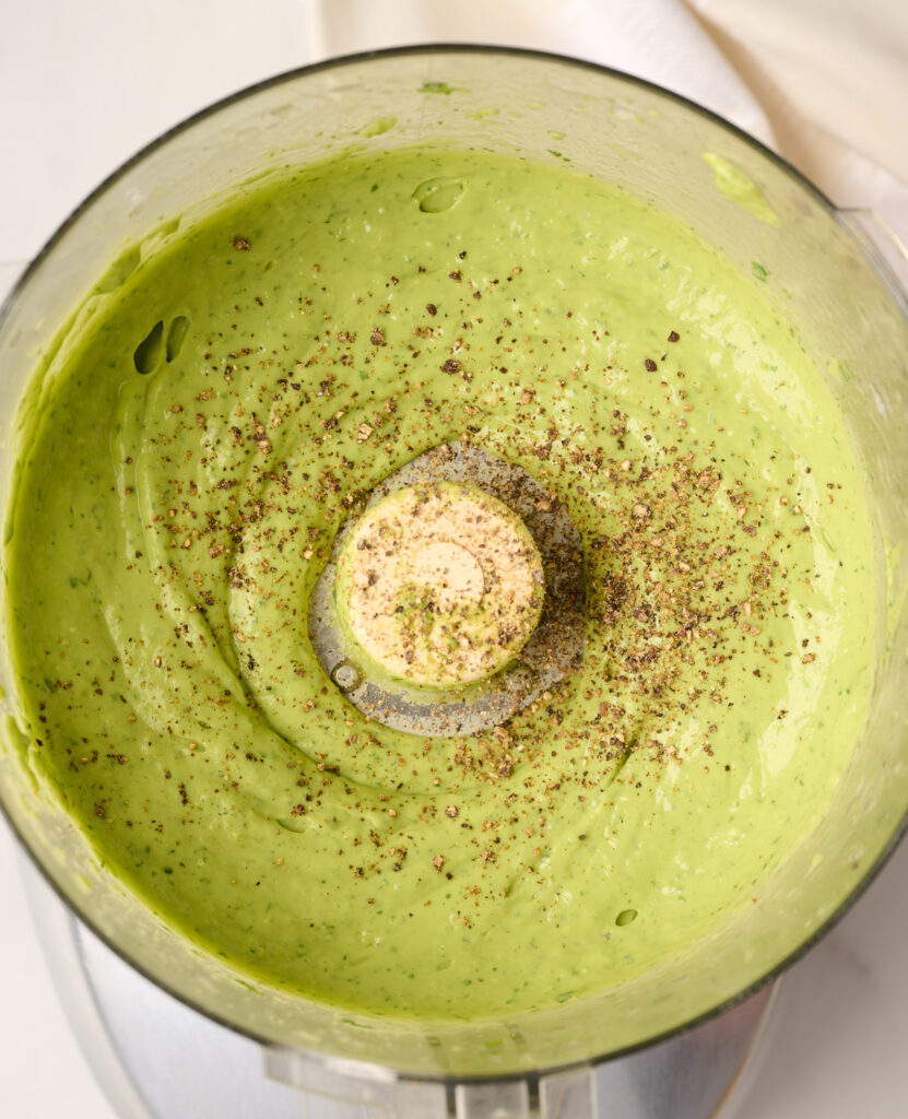 Black pepper added to finished avocado dressing inside a food processor, with white linen in the background.