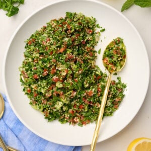 Half-eaten bowl of tabouli salad with a gold spoon, surrounded by lemon slices, herbs, and olive oil.