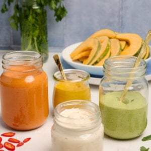 Finished tahini sauce in a jar with a gold spoon, alongside jars of red pepper tahini sauce, turmeric tahini sauce, and herb tahini sauce, with squash, herbs, peppers, and red onion in the background.