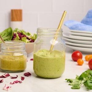 Finished avocado dressing in a jar with a gold spoon and a smaller jar of spicy avocado dressing with a gold spoon next to it, styled with a salad, pepper grinder, plates, blue linen, tomatoes, cilantro, and radicchio.