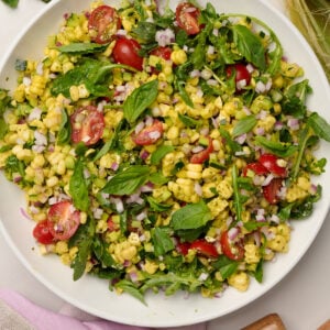 Close-up shot of fully composed corn salad with purple linen, wooden utensils, a corn cob, and basil in the background.