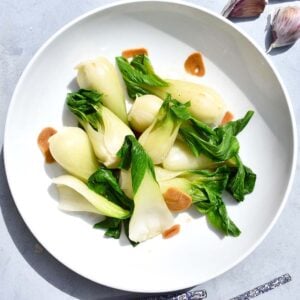 Plated baby bok choy with browned garlic in a white bowl, styled with chopsticks and garlic cloves.