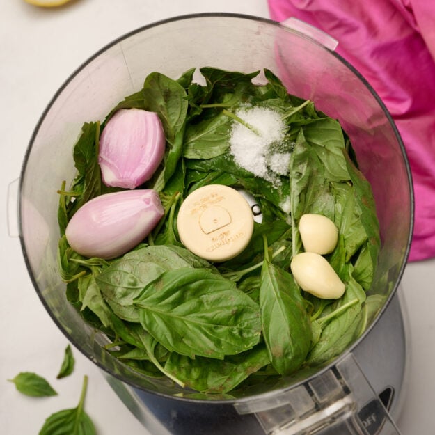 Basil, shallot, garlic, and salt in a food processor ready to be pulsed, with cut lemons, basil leaves