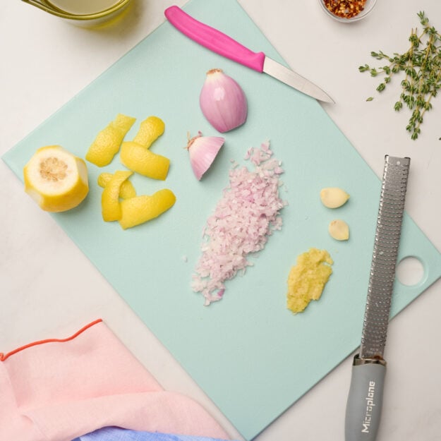 Minced shallots and garlic on a blue cutting board with lemon, lemon peel, a pink paring knife, and a Microplane, surrounded by thyme, chili flakes, olive oil, and pink and blue linens.