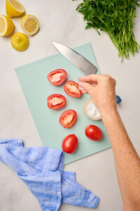 Hand cutting Roma tomatoes on a blue cutting board with salted tomatoes to remove moisture, parsley, lemons, and a chef’s knife on the side.