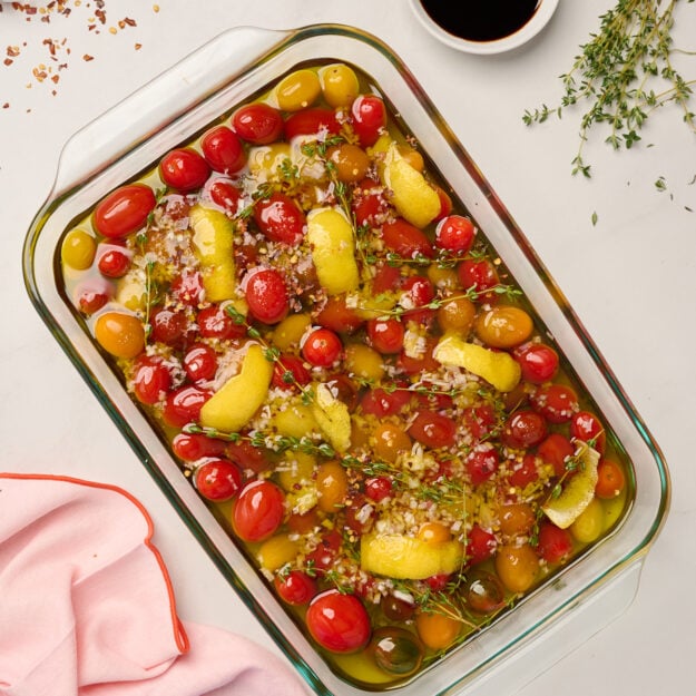 Glass baking dish filled with uncooked tomato confit ingredients, including tomatoes, olive oil, garlic, shallots, lemon peel, salt, chili flakes, and thyme, ready for the oven.