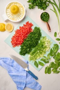 Chopped tomatoes, parsley, green onions, mint, and lemons on a blue cutting board with a lemon juicer.