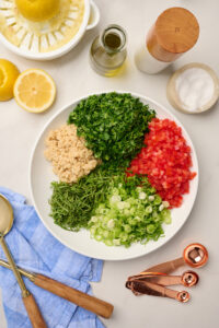 Large bowl with cooked bulgur, parsley, tomatoes, green onions, and mint with lemons juiced on the side, olive oil, salt, pepper, and serving utensils.