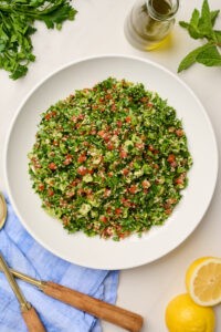 Overhead view of plated tabouli salad with lemon slices, gold and wooden serving utensils, blue linen, parsley, olive oil, and mint.