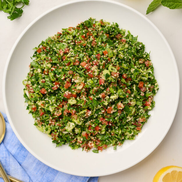 Overhead view of plated tabouli salad with lemon slices, gold and wooden serving utensils, blue linen, parsley, olive oil, and mint.