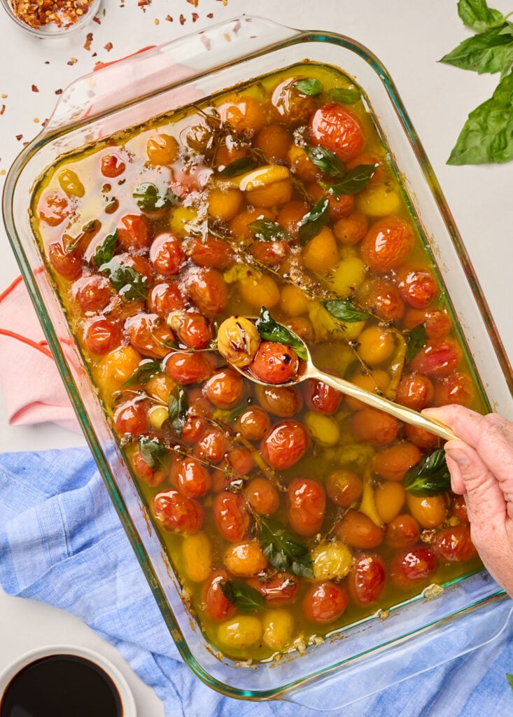 Hand holding a gold spoon of finished tomato confit over a glass baking dish, with basil leaves, chili flakes, and pink and blue linens in the background.