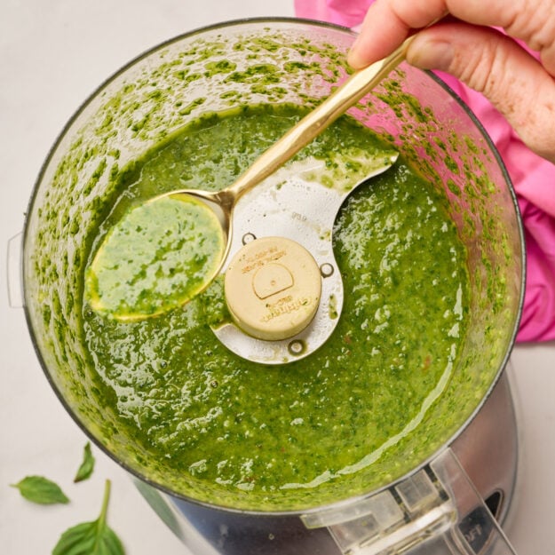 Hand holding a gold spoon filled with basil vinaigrette over a food processor, with basil leaves and pink linen in the background.
