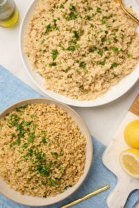 Close-up of cooked fine bulgur and coarse bulgur with gold spoon, blue linen, lemons, parsley, and olive oil.