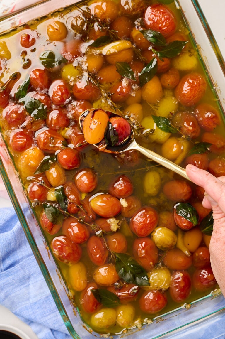 Hand holding a gold spoon of finished tomato confit drizzled with balsamic glaze, styled with pink and blue linens.