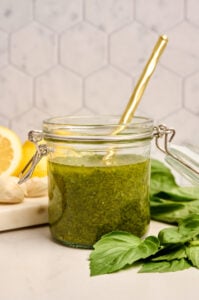 Finished jar of basil vinaigrette with a gold spoon inside, surrounded by fresh basil leaves, garlic, and lemon on a marble cutting board.