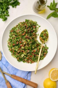 Half-eaten bowl of tabouli salad with a gold spoon, surrounded by lemon slices, herbs, and olive oil.