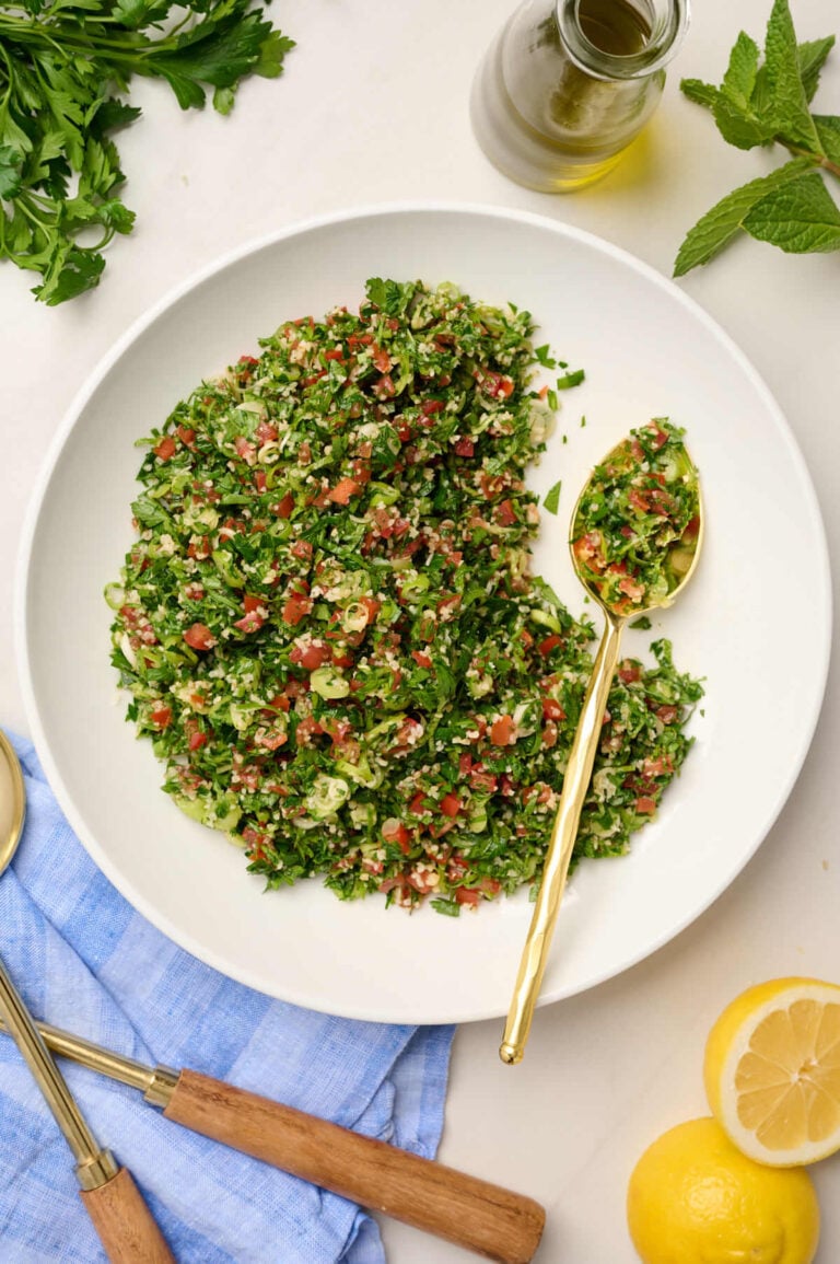 Half-eaten bowl of tabouli salad with a gold spoon, surrounded by lemon slices, herbs, and olive oil.