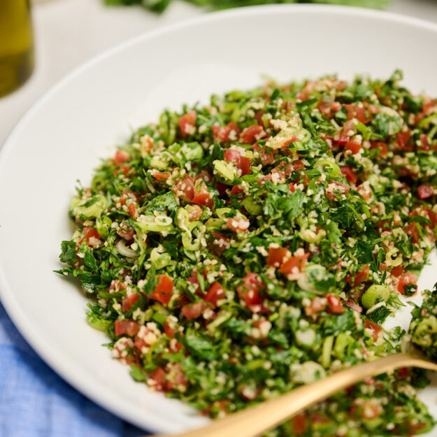 Close-up side view of tabouli salad with a gold spoon, blue linen, olive oil, and fresh herbs in the background.