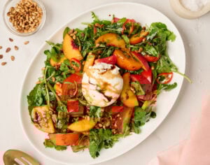 Close-up shot of plated burrata salad on an oval platter topped with a burrata ball, balsamic glaze, and toasted pine nuts, styled with peach linen and a gold serving spoon.