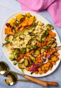 Serving platter of roasted vegetables on a blue tile surface with utensils and linen nearby