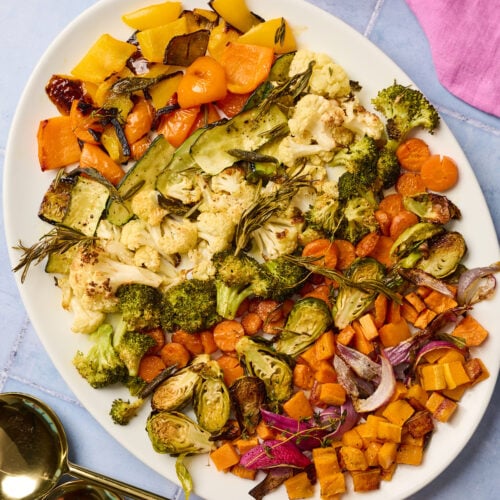 Serving platter of roasted vegetables on a blue tile surface with utensils and linen nearby