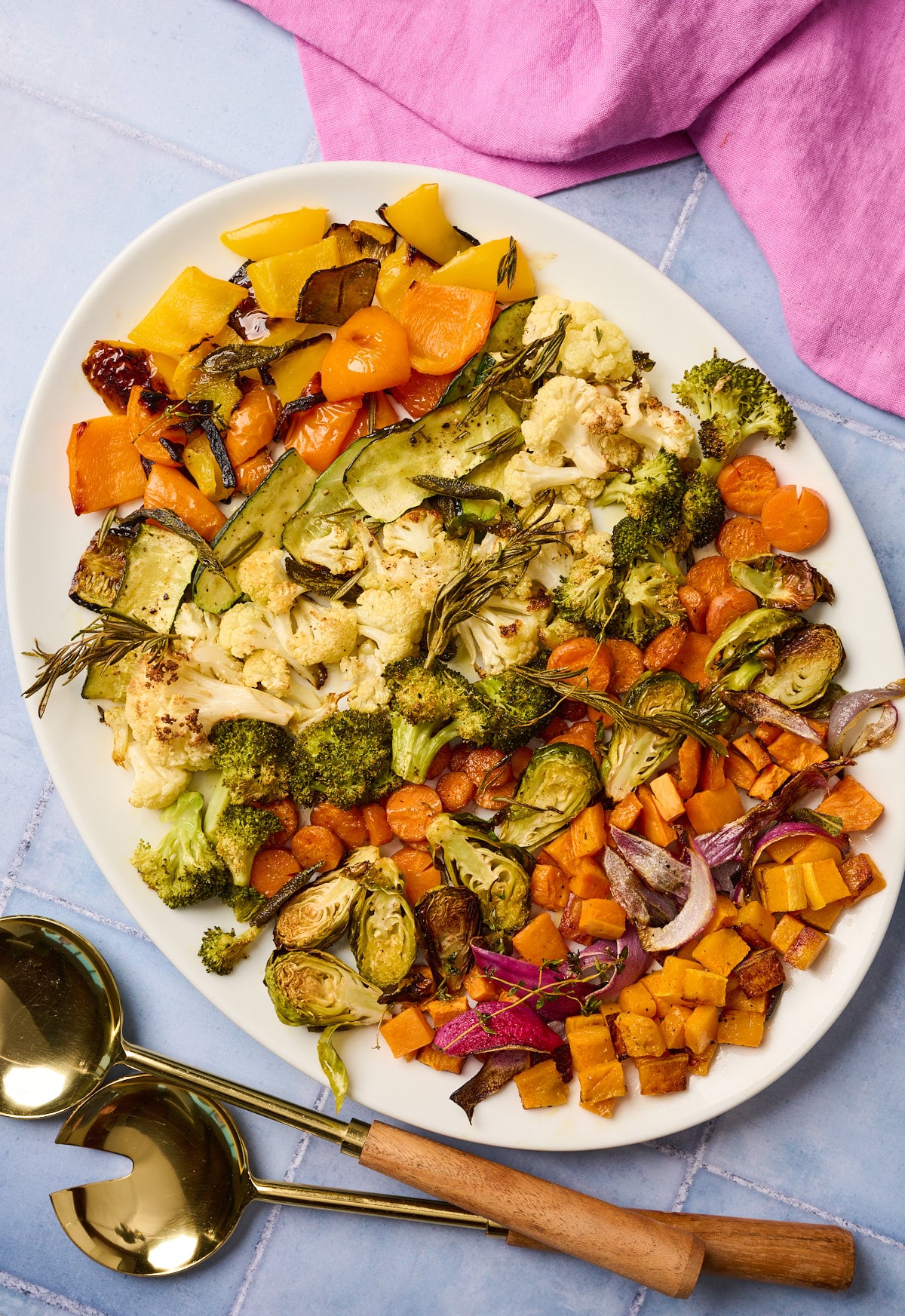Serving platter of roasted vegetables on a blue tile surface with utensils and linen nearby