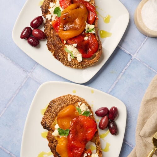 Two plates with roast meat topped with roasted bell peppers, crumbled feta, oregano sprigs, and black olives
