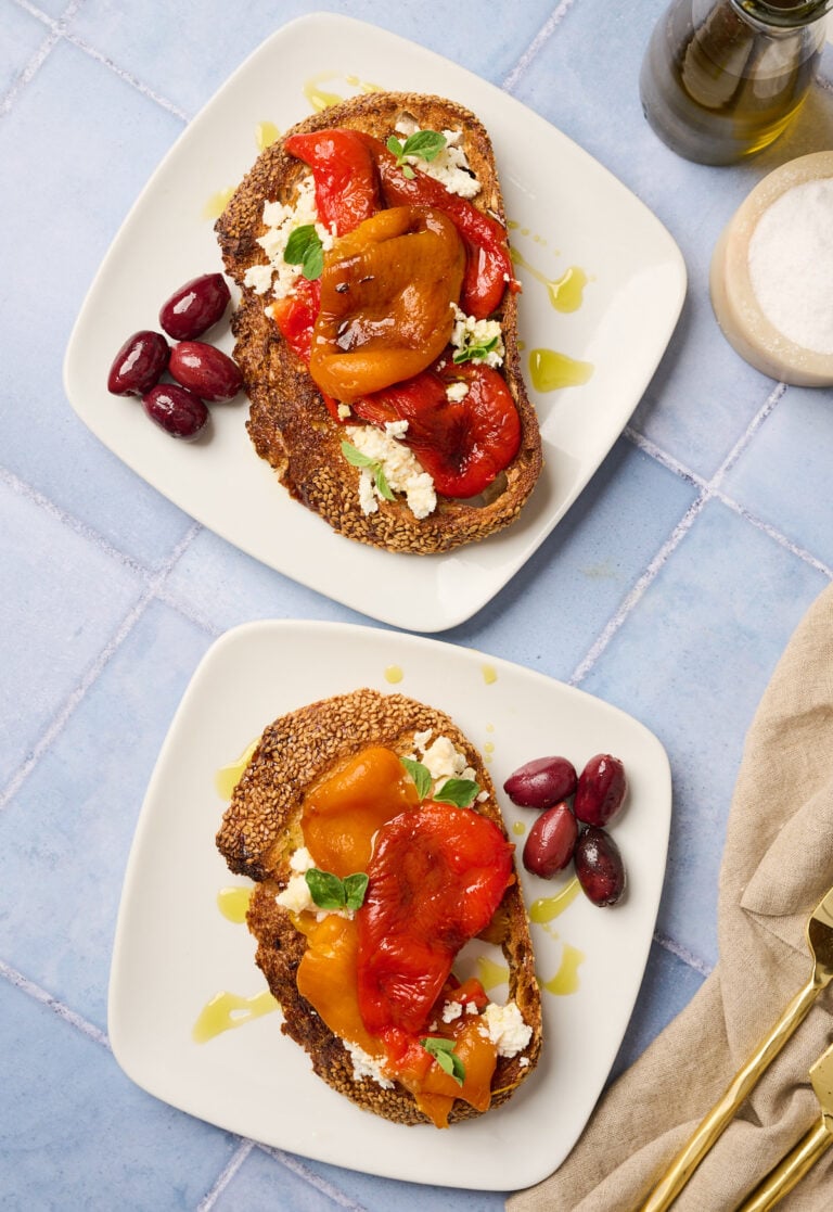 Two plates with roast meat topped with roasted bell peppers, crumbled feta, oregano sprigs, and black olives