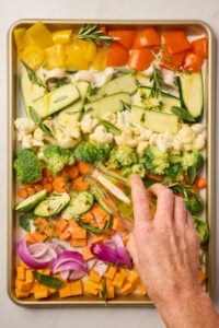 Sheet pan of raw vegetables arranged in a single layer with a hand drizzling olive oil