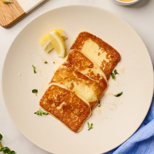 Pan-fried saganaki cheese served on a plate with honey drizzle and lemon wedges on white marble