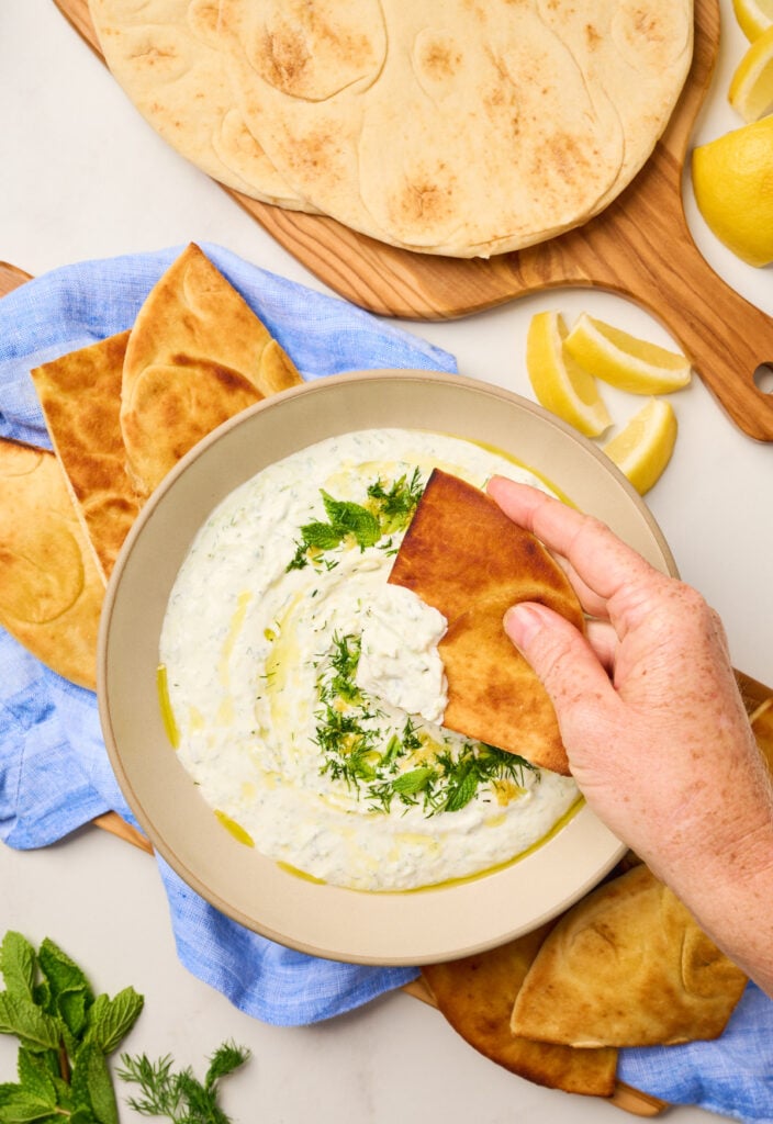 Hand dipping toasted pita into creamy tzatziki with olive oil and herbs